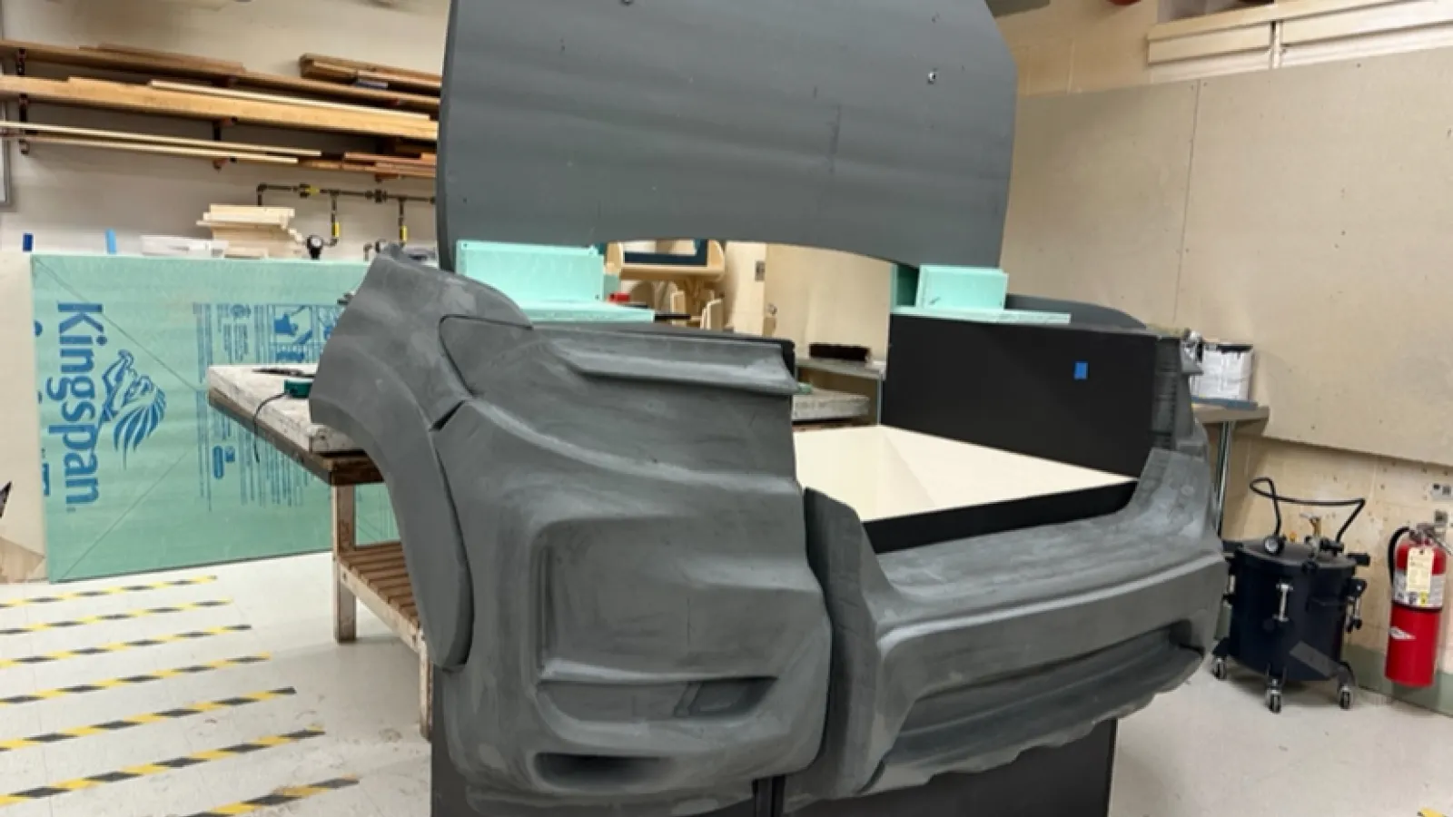 A model of the front part of a pickup truck (from in front of the side mirrors forward) sits in the CNC room in which it was assembled. The base of the model is made up of painted wooden boxes, and attached to this base are foam pieces that make up the shape of the truck, sanded smooth. The hood of the frunk is open, sitting on top of the model standing straight up. The grille is absent to show the frunk interior.
