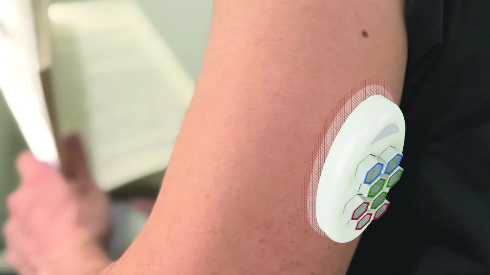 A wearable B.Well device attached to a person’s upper arm. The device has hexagonal, color-coded dosing cabins and is secured with a skin patch. The person is holding an open book, showing the device being used in a daily setting.