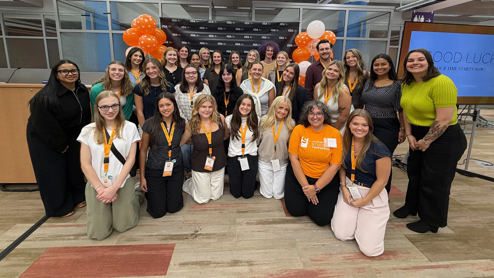 A large group of interior design students and a faculty member posing for a group photo at the IIDA Student Conference with orange balloon decorations and a display screen reading "GOOD LUCK".