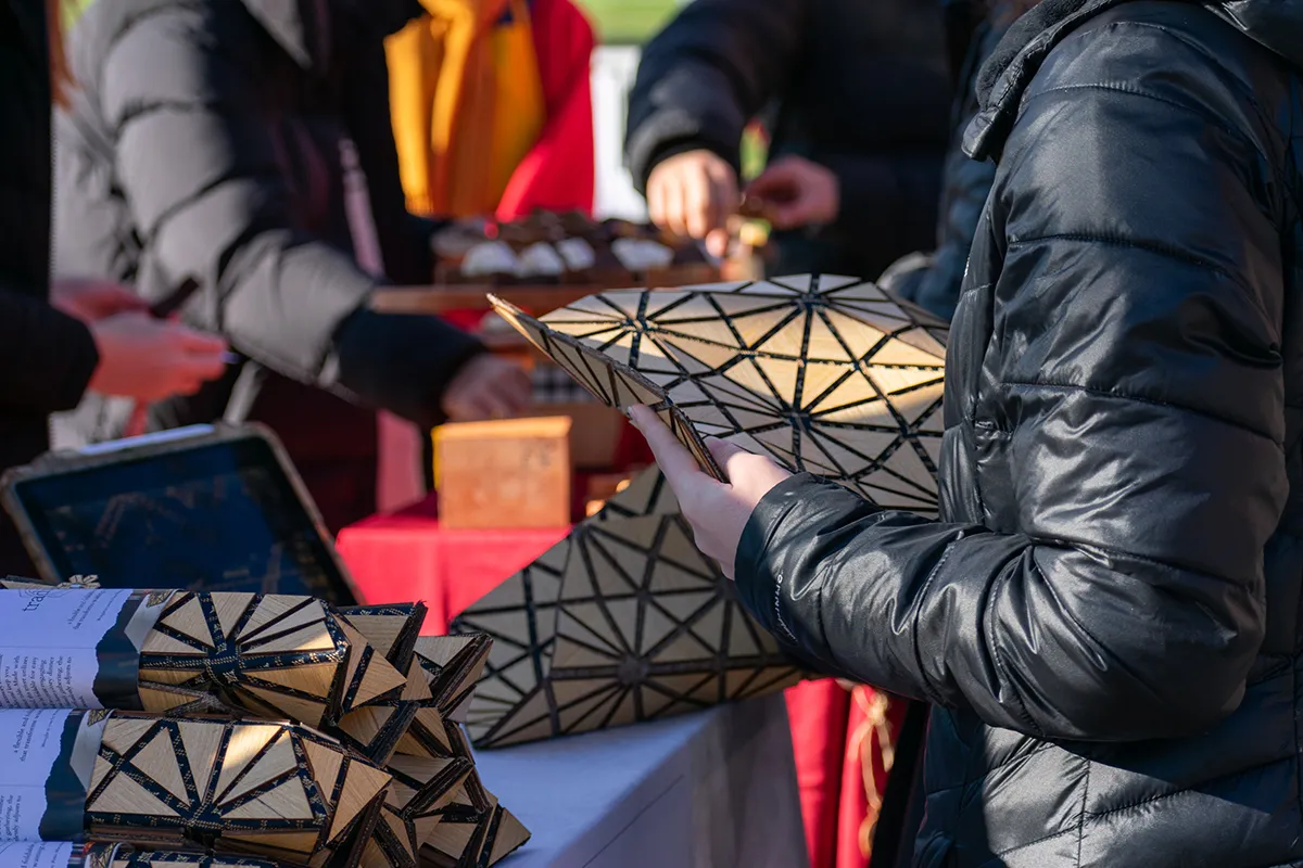 Several market guests handle home goods products over a market display table; the product in the foreground is a trivet made of tessellating, triangular wooden pieces and a dark-colored fabric.