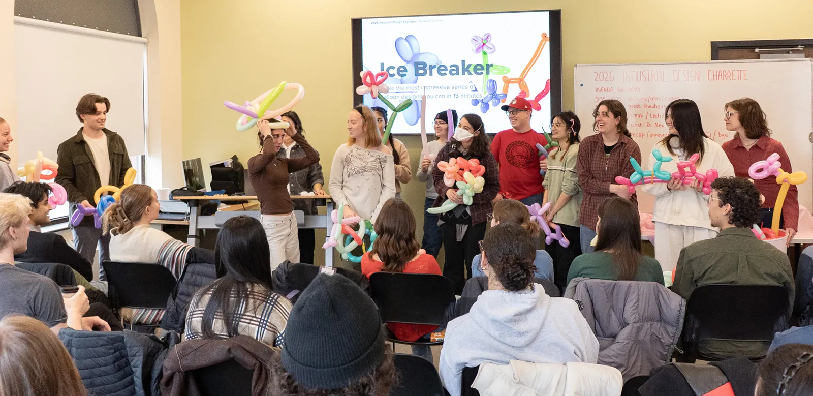 Design students standing at the front of a room in front of a presentation reading “Icebreaker”. The students are holding colorful balloon animal creations.