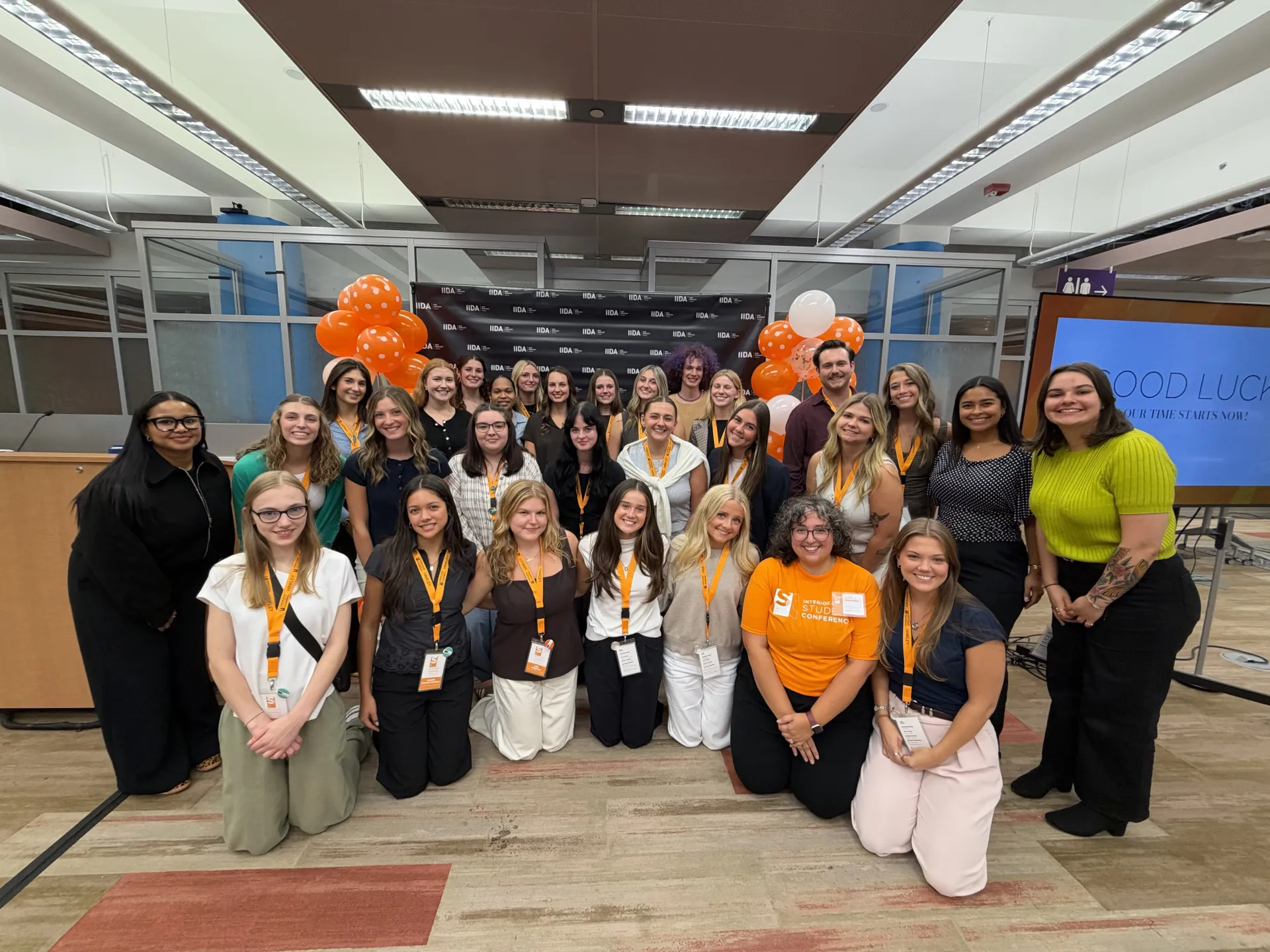 A large group of interior design students and a faculty member posing for a group photo at the IIDA Student Conference with orange balloon decorations and a display screen reading "GOOD LUCK".